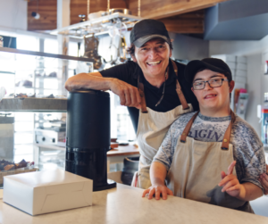 Pictured young man with down sydrome wearing a green shirt and apron working at a coffee shop with a woman support person behind him.  